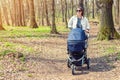 Beautiful young adult woman walking with baby in stroller through forest or park on bright sunny day. Healthy lifestyle and