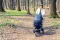 Beautiful young adult woman walking with baby in stroller through forest or park on bright sunny day. Healthy lifestyle and