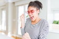 Beautiful young african american woman with afro hair wearing crazy pink heart glasses excited for success with arms raised