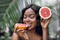 Beautiful young african american woman with grapefruit citrus fruit and palm leaves on background, looking pleasant and