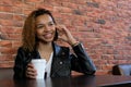 A beautiful young African-American woman in a jacket with a white paper glass in one hand, sitting at a table and smiling while ta Royalty Free Stock Photo