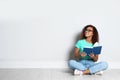 Beautiful young African-American woman reading book on wooden floor near light wall Royalty Free Stock Photo