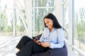 Beautiful young african woman looking at smart phone and smiling while sitting on bean bag chair with panoramic windows Royalty Free Stock Photo