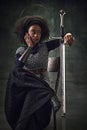 Beautiful young African woman, medieval warrior sitting on chair, leaning on sword against vintage green background Royalty Free Stock Photo