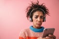 Beautiful young african woman posing isolated over pink wall background listening music with headphones using mobile phone Royalty Free Stock Photo