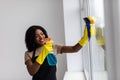 Beautiful young african woman is using a duster and a spray, looking at camera and smiling while cleaning windows in the house Royalty Free Stock Photo