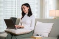 A beautiful Asian female is using her laptop on a sofa in her living room. Work from home Royalty Free Stock Photo