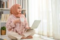 Beautiful young Asian Muslim woman sits on the living room's floor sipping coffee, using her laptop Royalty Free Stock Photo