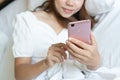 Beautiful young Asian smiling woman lying in white bed and using a phone in her bedroom. Technology, free time, communication
