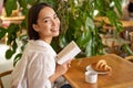 Beautiful young asian woman with a book in hands, sitting in cafe, drinking coffee and eating croissant, smiling Royalty Free Stock Photo