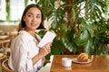 Beautiful young asian woman with a book in hands, sitting in cafe, drinking coffee and eating croissant, smiling Royalty Free Stock Photo