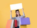 Beautiful young Asian woman carrying shopping bags and holding white banner with copy space isolated on yellow background Royalty Free Stock Photo
