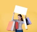 Beautiful young Asian woman carrying shopping bags and holding white banner with copy space isolated on yellow background Royalty Free Stock Photo