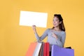 Beautiful young Asian woman carrying shopping bags and holding white banner with copy space isolated on yellow background Royalty Free Stock Photo