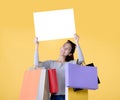 Beautiful young Asian woman carrying shopping bags and holding white banner with copy space isolated on yellow background Royalty Free Stock Photo