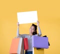 Beautiful young Asian woman carrying shopping bags and holding white banner with copy space isolated on yellow background Royalty Free Stock Photo