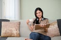 Beautiful young Asian woman sipping hot tea or coffee and reading a book on her sofa Royalty Free Stock Photo