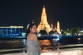 Beautiful young asian woman in dress standing and looking at camera on the deck with Wat Arun glowing in the night. Dinner cruise