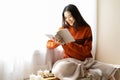 Beautiful young Asian woman reading book wearing knitted warm sweater sit on windowsill in room decorated with candle at home. Royalty Free Stock Photo