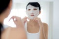 Beautiful young asian woman with sheet facial mask and looking mirror at home. Royalty Free Stock Photo