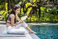 Beautiful young asian woman sits on the pool`s edge outdoors. She reading book while relaxing near swimming pool Royalty Free Stock Photo