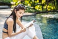 Beautiful young asian woman sits on the pool's edge outdoors. She reading book while relaxing near swimming pool Royalty Free Stock Photo