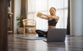 A beautiful young woman stretching arms while watching online workout tutorials on laptop at home