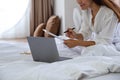 A beautiful young asian woman using laptop computer and working on paperwork while sitting on bed at home Royalty Free Stock Photo