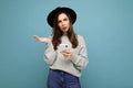 Beautiful young asking dissatisfied brunette woman wearing black hat and grey sweater holding smartphone looking at