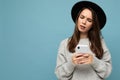 Beautiful young asking dissatisfied brunette woman wearing black hat and grey sweater holding smartphone looking down