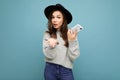 Beautiful young asking dissatisfied brunette woman wearing black hat and grey sweater holding smartphone looking at