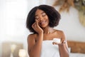 Beautiful young black woman applying face cream from jar, wearing bath towel, enjoying spa procedure at home Royalty Free Stock Photo