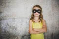 Blond girl with black superhero mask and yellow shirt is posing in front of concrete background