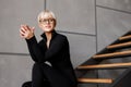 A beautiful young blond girl in stylish glasses and a tight black suit is sitting on the stairs and looking away. Fashion Royalty Free Stock Photo