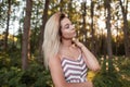 Beautiful young blond woman in a glamorous pink striped sundress relaxes standing in the forest on a sunny hot summer day.