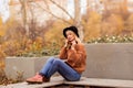 A beautiful young blonde in a brown warm sweater, black felt hat, blue jeans and boots sits on a bench in the autumn in the park, Royalty Free Stock Photo