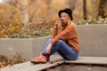 Beautiful young blonde in a brown warm sweater, black felt hat, blue jeans and boots sits on a bench in the autumn in the park, Royalty Free Stock Photo