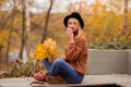 Beautiful young blonde in a brown warm sweater, black felt hat, blue jeans and boots sits on a bench in the autumn in the park, Royalty Free Stock Photo