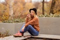 Beautiful young blonde in a brown warm sweater, black felt hat, blue jeans and boots sits on a bench in the autumn in the park, Royalty Free Stock Photo