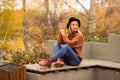 Beautiful young blonde in a brown warm sweater, black felt hat, blue jeans and boots sits on a bench in the autumn in the park, Royalty Free Stock Photo