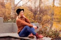 Beautiful young blonde in a brown warm sweater, black felt hat, blue jeans and boots sits on a bench in the autumn in the park, Royalty Free Stock Photo