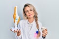 Beautiful young blonde doctor woman holding anatomical model of knee joint and protein powder relaxed with serious expression on