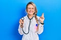 Beautiful young blonde doctor woman holding stethoscope gesturing finger crossed smiling with hope and eyes closed Royalty Free Stock Photo