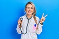 Beautiful young blonde doctor woman holding stethoscope smiling looking to the camera showing fingers doing victory sign Royalty Free Stock Photo