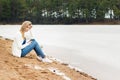 Beautiful young blonde girl in jeans and a white shirt sitting on the shore of the frozen cold of the lake near the forest Royalty Free Stock Photo