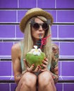 Beautiful young blonde hipster in a straw hat and sunglasses sitting on the purple stairs with coconut in hand. Rain