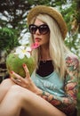 Beautiful young blonde hipster in a straw hat and sunglasses sitting in a tropical garden with coconut in hand. Rain