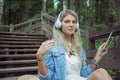 Beautiful young blonde hipster woman listening music in headphones in park with tablet in hands, sitting on wooden steps Royalty Free Stock Photo