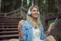 Beautiful young blonde hipster woman listening music in headphones in park with tablet in hands, sitting on wooden steps Royalty Free Stock Photo