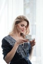 Beautiful young blonde woman in a blue robe by the window. Drinks coffee or tea from a white cup with a saucer. Morning Royalty Free Stock Photo